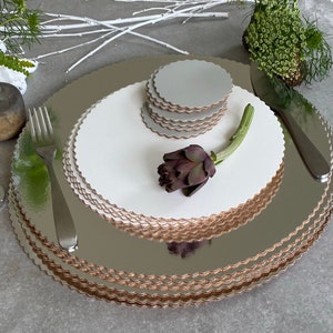 Puede incluir: Una pila de cartones para pasteles y posavasos plateados y blancos con bordes festoneados. Una alcachofa morada oscura y cubiertos están dispuestos sobre los cartones. Los cartones tienen un brillo metálico y los posavasos están apilados.
