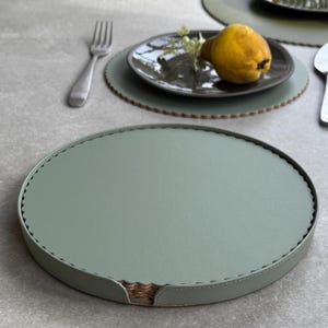 May include: Sage green placemats with a scalloped edge and stitched detail. A stack of matching coasters is visible. A plate with a pear and sprig of greenery sits on top of a placemat. A fork and spoon are also visible.