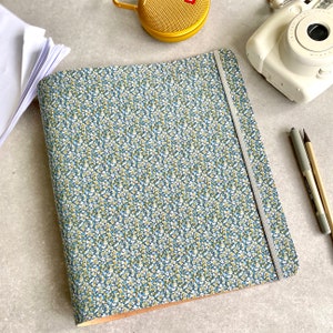 May include: A notebook with a floral pattern in blue, white, and yellow. It features a grey elastic closure. Other items include a white camera, gold speaker, pens, and a stack of paper. The notebook is likely A4 size (approx. 21 x 29.7 cm).