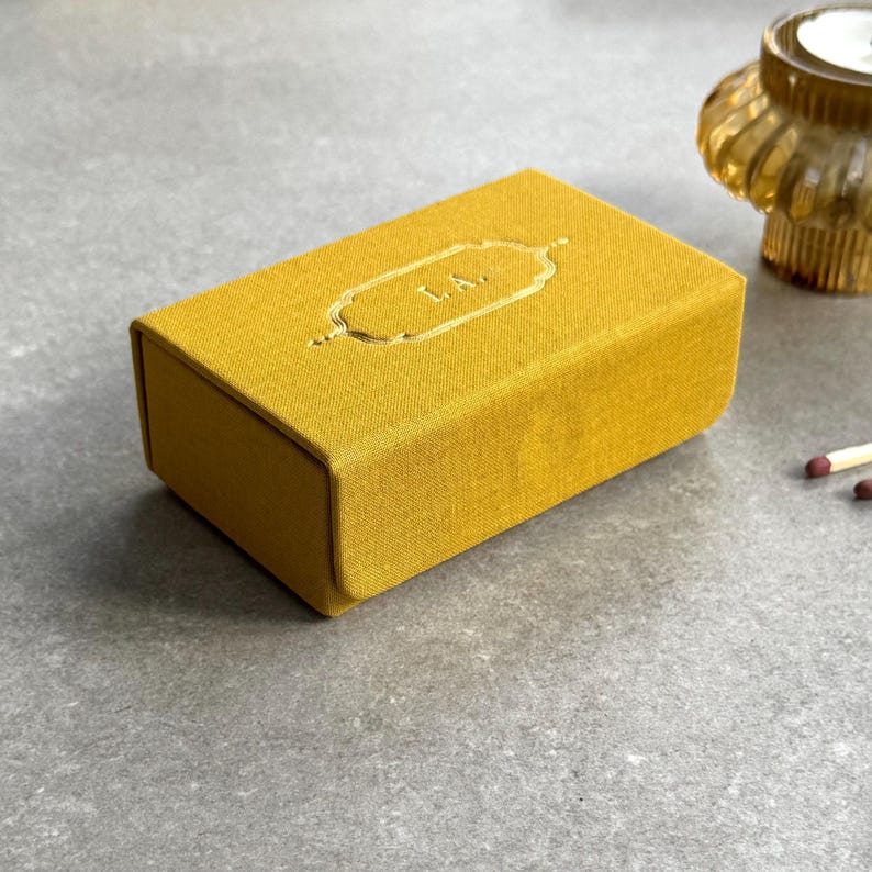 May include: Mustard yellow rectangular box with gold embossed detailing and the letters "L.A." on the top. The box is made of a textured fabric. A small glass candle holder and matches are in the background.