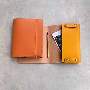 Peut inclure: Un carnet en cuir orange avec un élastique, un crayon doré et un plateau en cuir jaune avec des boutons-pression argentés. Une petite carte avec le texte Wild Chicory est glissée dans le rabat en cuir ouvert reliant le carnet et le plateau.