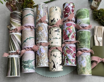 May include: An assortment of decorative crackers with marbled patterns in shades of green, purple, and cream. Each cracker is adorned with a red and white striped ribbon. The crackers are displayed on a pale green surface.