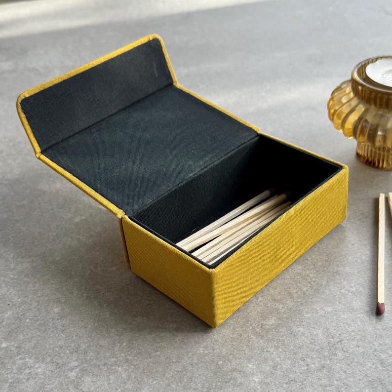 May include: Mustard-yellow matchbox with a black interior, containing wooden matches. The rectangular box has a hinged lid. A small candle holder is visible in the background.