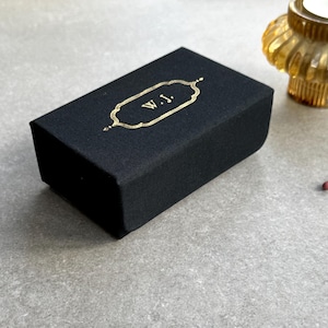May include: A black rectangular box with gold lettering that reads "W.J." The box has a gold decorative border. The box is on a grey surface. A gold candle holder is in the background.