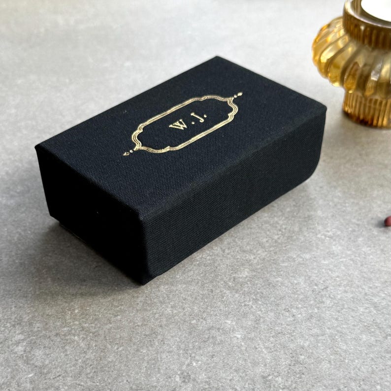 May include: A black rectangular box with gold lettering that reads "W.J." The box has a gold decorative border. The box is on a grey surface. A gold candle holder is in the background.