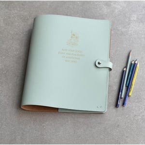 May include: A light green leather portfolio with a snap closure and brown interior. The cover features a gold logo and the text: ADD YOUR LOGO FONT AND PLACEMENT OF ADDITIONAL TEXT HERE. Four colored pencils are positioned to the right of the portfolio.