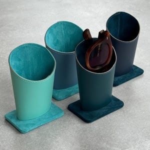 Peut inclure: Quatre étuis à lunettes en cuir dans les tons de turquoise, de bleu et de marine. Chaque étui est cylindrique avec une base carrée et un intérieur contrastant. Un étui contient une paire de lunettes de soleil marron.