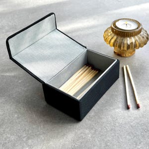 May include: A black matchbox with a grey interior, open to show a bundle of matches. Two matches are placed outside the box. An amber-coloured candle holder with a lit candle is also visible.