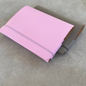 Könnte beinhalten: Zwei Notizbücher, eines in hellem Lila und eines in Grau, sind auf einer grauen Oberfläche gestapelt. Jedes Notizbuch hat einen passenden Gummibandverschluss. Das hellviolette Notizbuch liegt oben und verdeckt teilweise das graue Notizbuch.