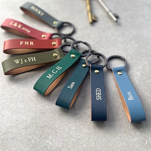 Puede incluir: Un juego de ocho llaveros de cuero con nombres e iniciales personalizados. Los llaveros son de varios colores, incluyendo verde, azul, rojo y marrón. Cada llavero tiene un anillo de metal.