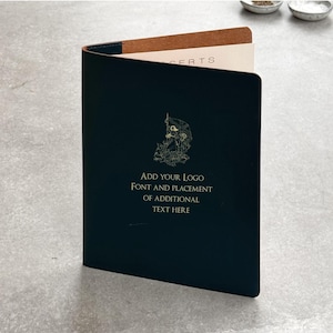 Op de afbeelding: Een donkerblauw leren boekje met gouden letters en een decoratief embleem. De tekst op de omslag luidt "ADD YOUR LOGO, FONT AND PLACEMENT OF ADDITIONAL TEXT HERE". De binnenpagina's zijn lichtbruin.