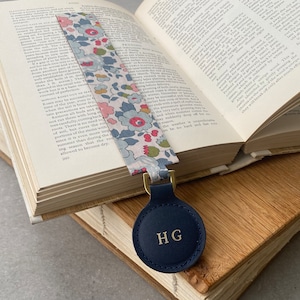 Può includere: Un segnalibro in pelle blu con un fiocco di metallo dorato e un'etichetta rotonda in pelle blu scuro con le iniziali "HG" in rilievo in bianco.