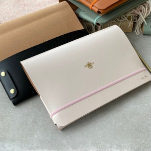 May include: A light pink leather notebook with a gold bee design and a pink elastic band closure. The notebook has the initials "SLM" embossed on the cover.