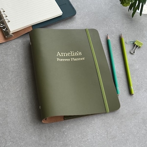 Può includere: Un'agenda verde oliva con la scritta "Amelia's Forever Planner" è mostrata. Un elastico verde tiene chiusa l'agenda. Due matite e una graffetta sono accanto all'agenda. Un taccuino aperto parzialmente visibile è sullo sfondo.