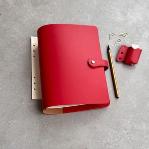 Może przedstawiać: Jasnoczerwony skórzany planer z zapięciem na zatrzask, zawierający papier w linie z otworami. Obok planera leży ołówek w kolorze złotym, dwa srebrne metalowe spinacze oraz mały czerwony skórzany dodatek z trzema okrągłymi wycięciami.