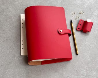 Peut inclure: Une couverture de carnet en cuir rouge vif avec une fermeture à pression, comprenant un porte-stylo rouge assorti, un stylo doré et deux trombones en métal argenté. Le carnet présente des feuilles lignées visibles sur le côté gauche.