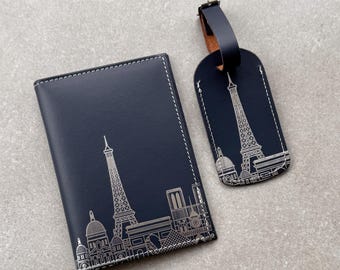 Peut inclure: Un porte-passeport bleu marine foncé et une étiquette de bagage assortie, tous deux ornés d'une illustration au trait blanc de la ligne d'horizon de Paris, incluant la tour Eiffel. Chaque article présente des coutures blanches sur les bords.