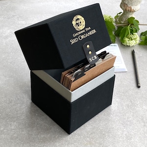 May include: Black seed organizer box with a gold lion logo and the text "Gardening Year Seed Organiser". The box is open and has brown card dividers labeled with months of the year.