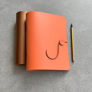 May include: An orange leather journal with a brown leather strap closure and a gold pencil. The journal is next to a rolled-up brown leather item. The items are on a gray surface.