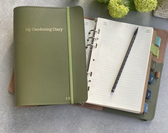 May include: A green leather bound journal with the words "My Gardening Diary" embossed on the front cover. The journal is open to a blank page with a pencil lying on top. The journal has a ribbon bookmark and a metal ring binder.