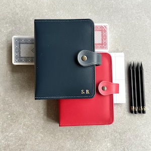 May include: Two leather wallets, one navy blue and one red, with snap closures. The navy wallet has gold initials "S.B." and is next to a deck of cards and a notepad. Five black pencils are also in the image.