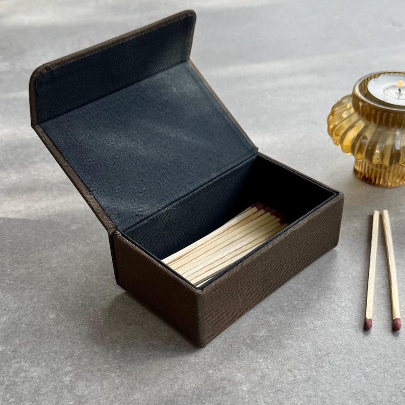 May include: A brown matchbox with a hinged lid, open to show a stack of matches. The interior is lined with black fabric. Two matches with red tips are placed beside the box. An amber-coloured candle holder is in the background.