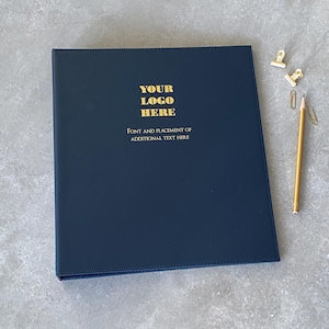 May include: A dark navy blue binder with gold foil text that reads YOUR LOGO HERE and FONT AND PLACEMENT OF ADDITIONAL TEXT HERE. The binder is accompanied by a gold pencil and two gold metal binder clips positioned to the right side.