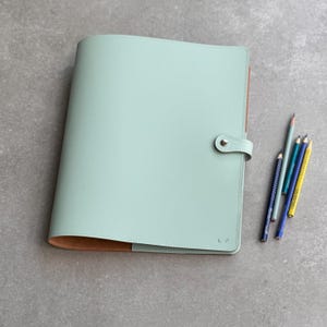 May include: A mint green folio with a tan interior, a snap closure, and the initials K.P. embossed on the front cover. Four sharpened pencils in shades of grey, teal, blue, and yellow are positioned to the right of the closed folio.