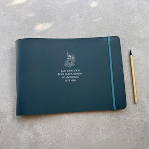 Op de afbeelding: Een donkerblauwgroen horizontaal notitieboek met een blauwgroene elastische sluitband en een geel potlood. De kaft bevat een witte lijntekening van een vogel en de tekst: ADD YOUR LOGO FONT AND PLACEMENT OF ADDITIONAL TEXT HERE.