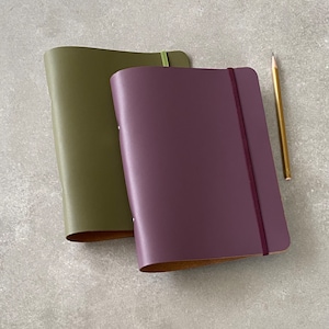 Puede incluir: Dos diarios encuadernados en cuero, uno verde y otro morado, con un lápiz sobre una superficie gris.