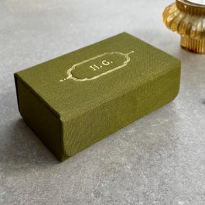 May include: An olive green rectangular box with gold-coloured detailing and the initials "H.G." in gold. The box has a textured fabric finish and a hinged lid. The background is a light grey surface.