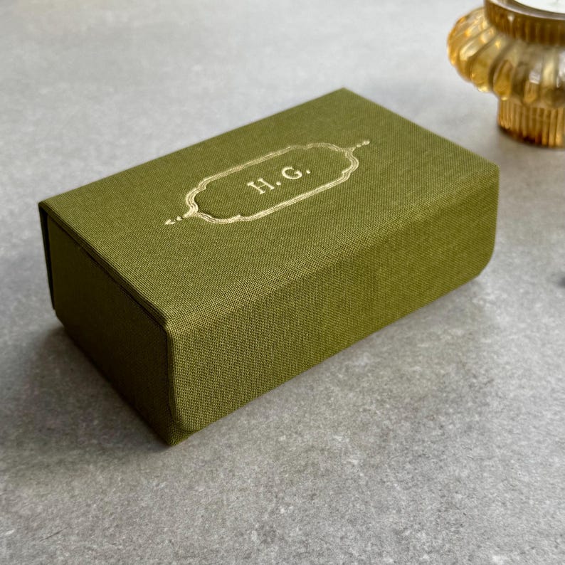 May include: An olive green rectangular box with gold-coloured detailing and the initials "H.G." in gold. The box has a textured fabric finish and a hinged lid. The background is a light grey surface.