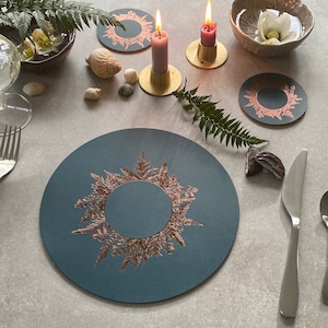 Könnte beinhalten: Ein Set mit vier runden Platzsets in Blau und Kupfer mit einem Farnmuster. Die Platzsets befinden sich auf einem weißen Tisch mit Kerzen und Muscheln.