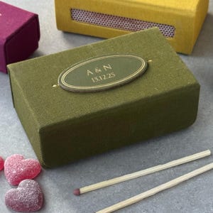 Peut inclure: Une boîte d'allumettes vert olive avec une étiquette ovale bordée d'or portant l'inscription "A & N 15.12.25". La boîte rectangulaire a une finition en tissu texturé. Deux allumettes et des bonbons en forme de cœur sont également visibles.