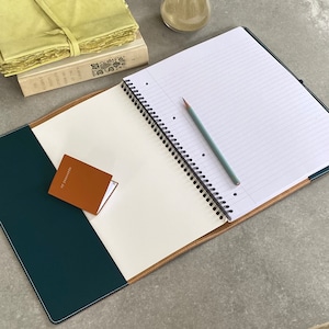 Może przedstawiać: Zielony skórzany notes z spiralnym oprawieniem i liniowanymi stronami. Mały brązowy skórzany notes z nadrukowanym słowem "remarkable" włożony do przedniej okładki. Jasnoniebieski ołówek leży na otwartych stronach.