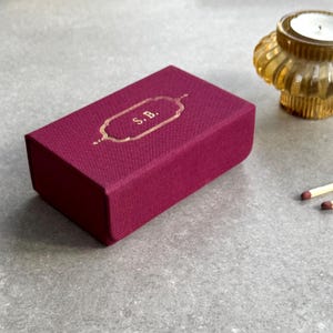 May include: A rectangular burgundy box with gold lettering that reads "S.B." is the focal point. A gold-coloured candle holder and two matches are also visible. The box has a textured surface and a decorative gold border.