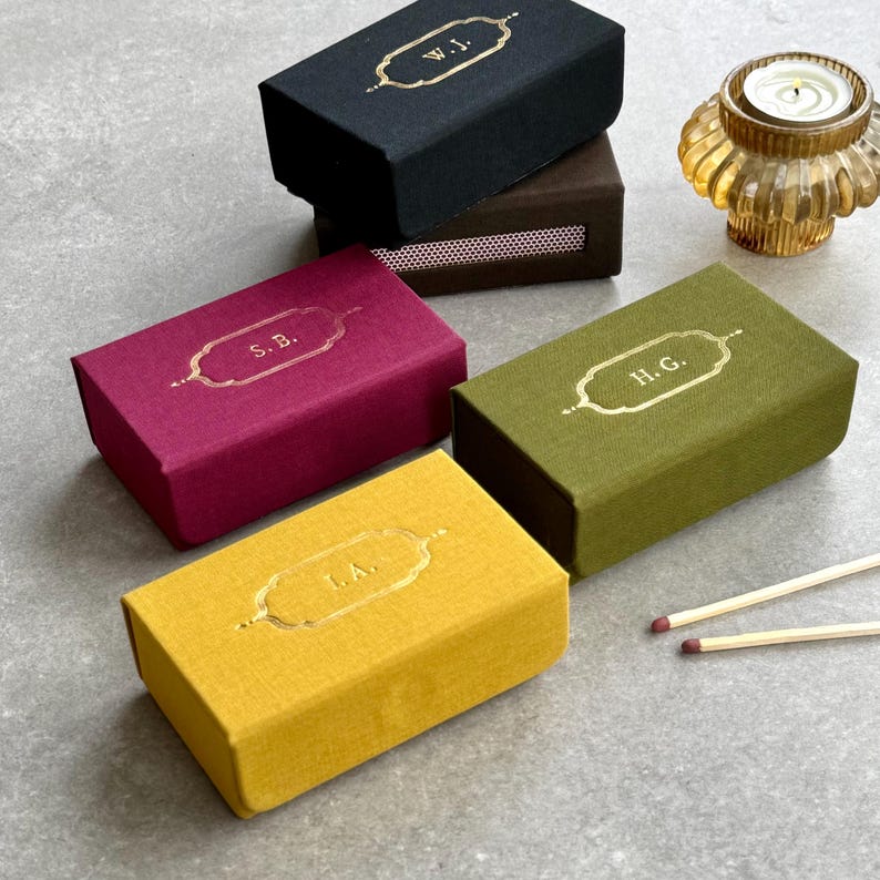 May include: A selection of rectangular matchboxes in black, brown, burgundy, olive green, and yellow. Each box has gold lettering with initials. A small candle holder and matches are also visible.