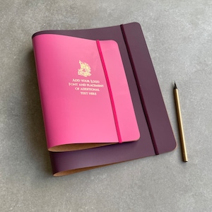 May include: A magenta notebook with a matching elastic band rests atop a larger, plum-colored notebook. The magenta notebook features gold text and a logo. A gold pencil lies to the right.