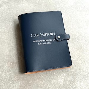 May include: A navy blue leather car history binder with the text "CAR HISTORY" and "1968 FORD MUSTANG GT REG: ABC 123D" in silver lettering. The binder has a snap closure.