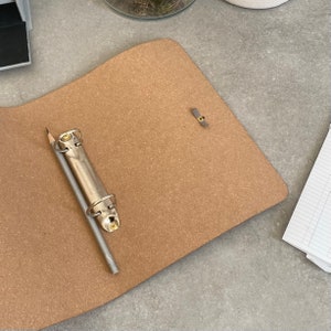 May include: A brown corkboard binder with a silver binder ring and a pencil. The binder has a gray metal closure.