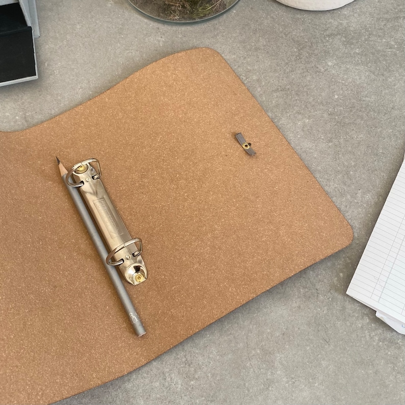 May include: A brown corkboard binder with a silver binder ring and a pencil. The binder has a gray metal closure.