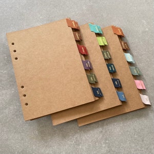 May include: Three brown paper notebooks with leather tabs for each month of the year. The tabs are in different colours, including red, green, blue, and purple.