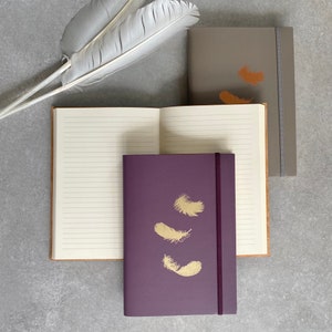 Könnte beinhalten: Drei Notizbücher mit verschiedenen farbigen Umschlägen. Die Notizbücher haben ein goldenes Federdesign auf der Vorderseite. Die Notizbücher liegen auf einer grauen Oberfläche.