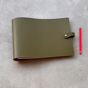 May include: A rectangular olive green notebook with a smooth finish and a side flap closure secured by a silver metal snap button. A bright red pencil is positioned vertically to the right of the notebook on a light gray textured surface.