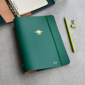 Może przedstawiać: Zielony, skórzany notes z motywem złotej pszczoły i inicjałami "AP" na okładce. Notes ma zielony, elastyczny pasek zamykający i leży na szarej powierzchni.