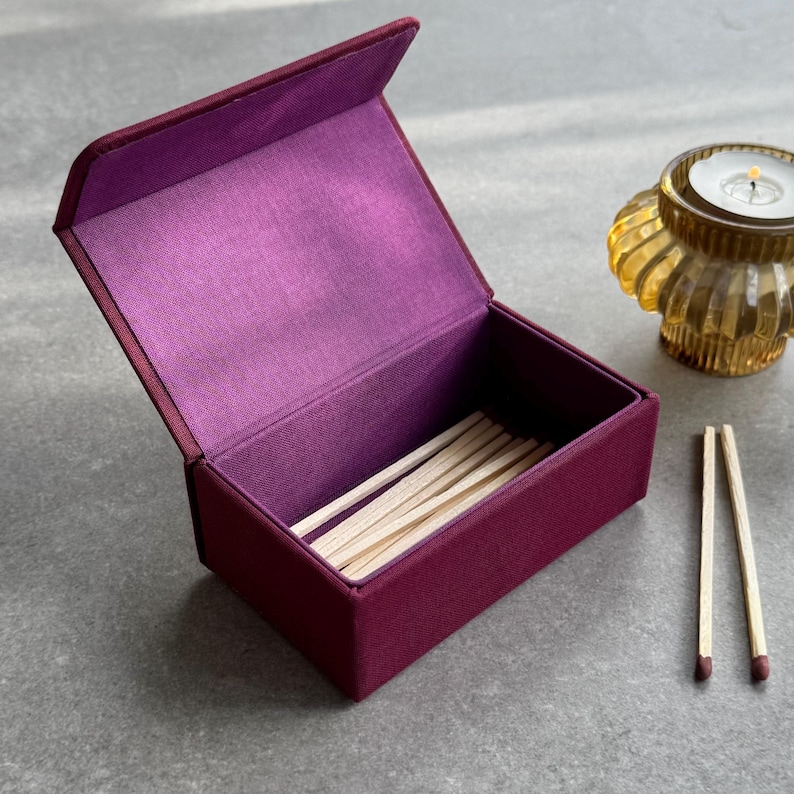 May include: A burgundy fabric matchbox, open to display wooden matches. Two matches with red tips are placed beside the box. A gold-coloured candle holder with a lit candle is in the background.