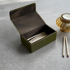 May include: An olive green matchbox with a hinged lid, open to reveal several matches. The interior is a dark brown. Two matches are placed outside the box. A small, amber-coloured candle holder is in the background.