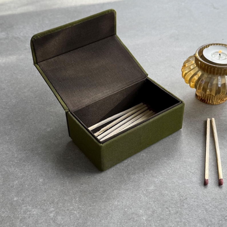 May include: An olive green matchbox with a hinged lid, open to reveal several matches. The interior is a dark brown. Two matches are placed outside the box. A small, amber-coloured candle holder is in the background.