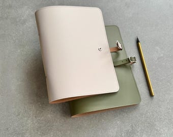 Peut inclure: Deux journaux en cuir, un crème et un vert olive, sont présentés. Le journal crème a une fermeture à bouton, et le journal vert olive a une fermeture à boucle. Un stylo doré est posé à côté.