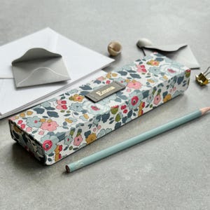 Puede incluir: Un estuche rectangular con un estampado floral en rosa, azul y rojo. El estuche tiene una etiqueta de cuero gris con el nombre "Emma". Un lápiz azul claro y artículos de papelería están al lado.